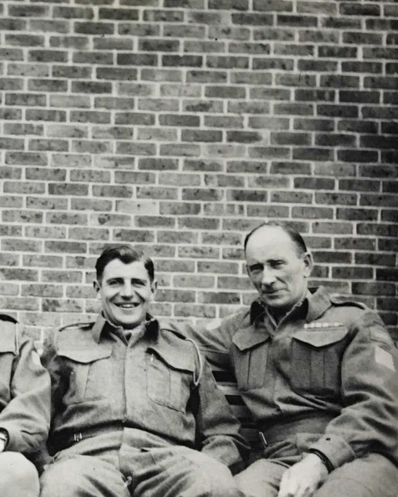 Photographie en noir et blanc – Archie, à droite, est assis sur un banc avec un homme beaucoup plus jeune. Les deux sont en uniforme. Ils sourient.