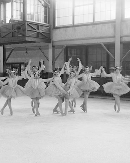 Une photographie en noir et blanc de dix patineurs en costume de tutu dans diverses poses. Le groupe est dans une patinoire couverte.